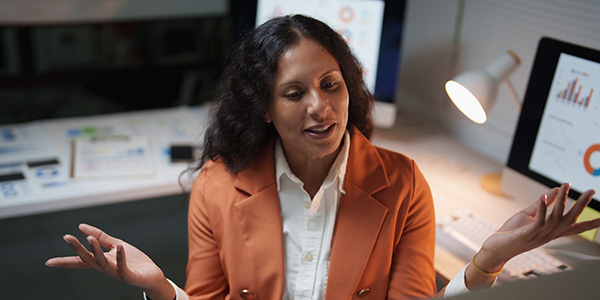 A woman in a professional blazer speaking and gesturing during a virtual meeting in an office setting.